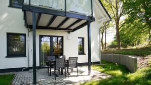 Une maison moderne dotée d’un patio couvert, d’un coin repas avec des chaises et une table, et offrant une vue sur la verdure environnante.