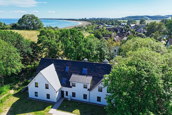 A white house with a dark roof, surrounded by lush greenery and a view of the sea.