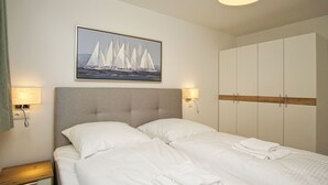 Un lit bien fait, avec du linge de lit blanc, une tête de lit et un tableau représentant des voiliers accroché au mur.