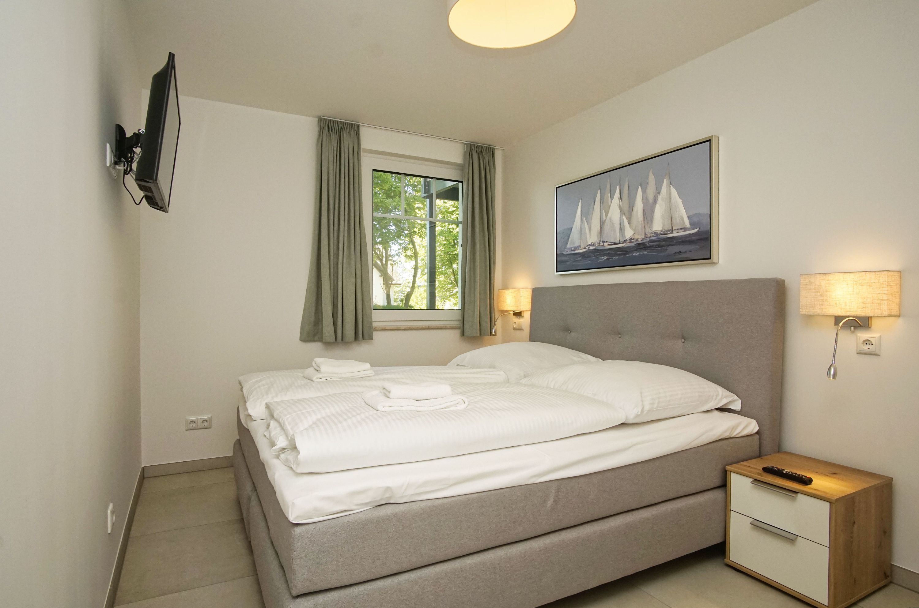 A neatly made bed with white linens, a bedside table with a lamp, a TV mounted on the wall, and a painting of sailboats above the bed.