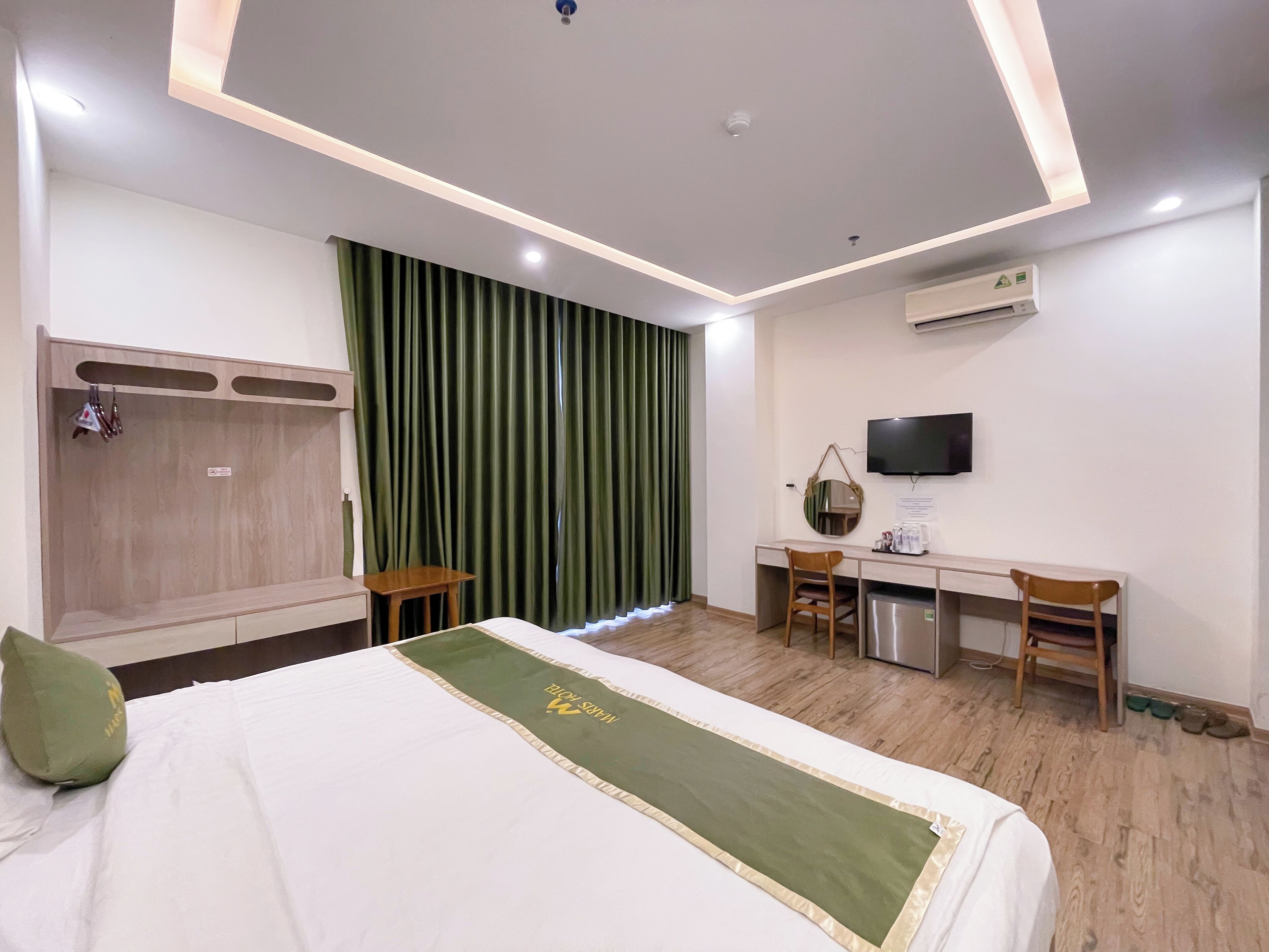 A hotel room with a bed, a desk, a chair, a TV, and an air conditioning unit.