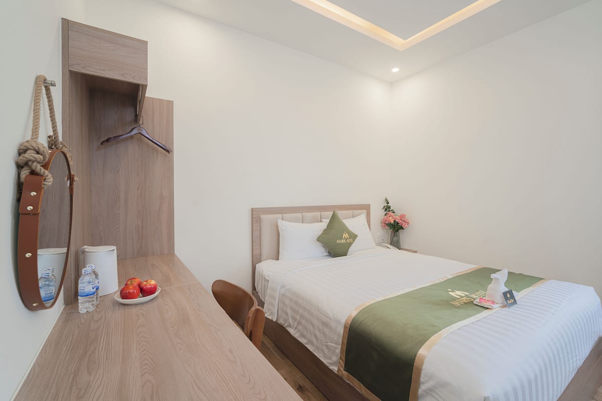 Ein ordentlich eingerichtetes Hotelzimmer mit einem Bett, einem Holztisch, einem Spiegel, einem aufgehängten Handtuch und einem kleinen Tisch mit Äpfeln und einer Wasserflasche.