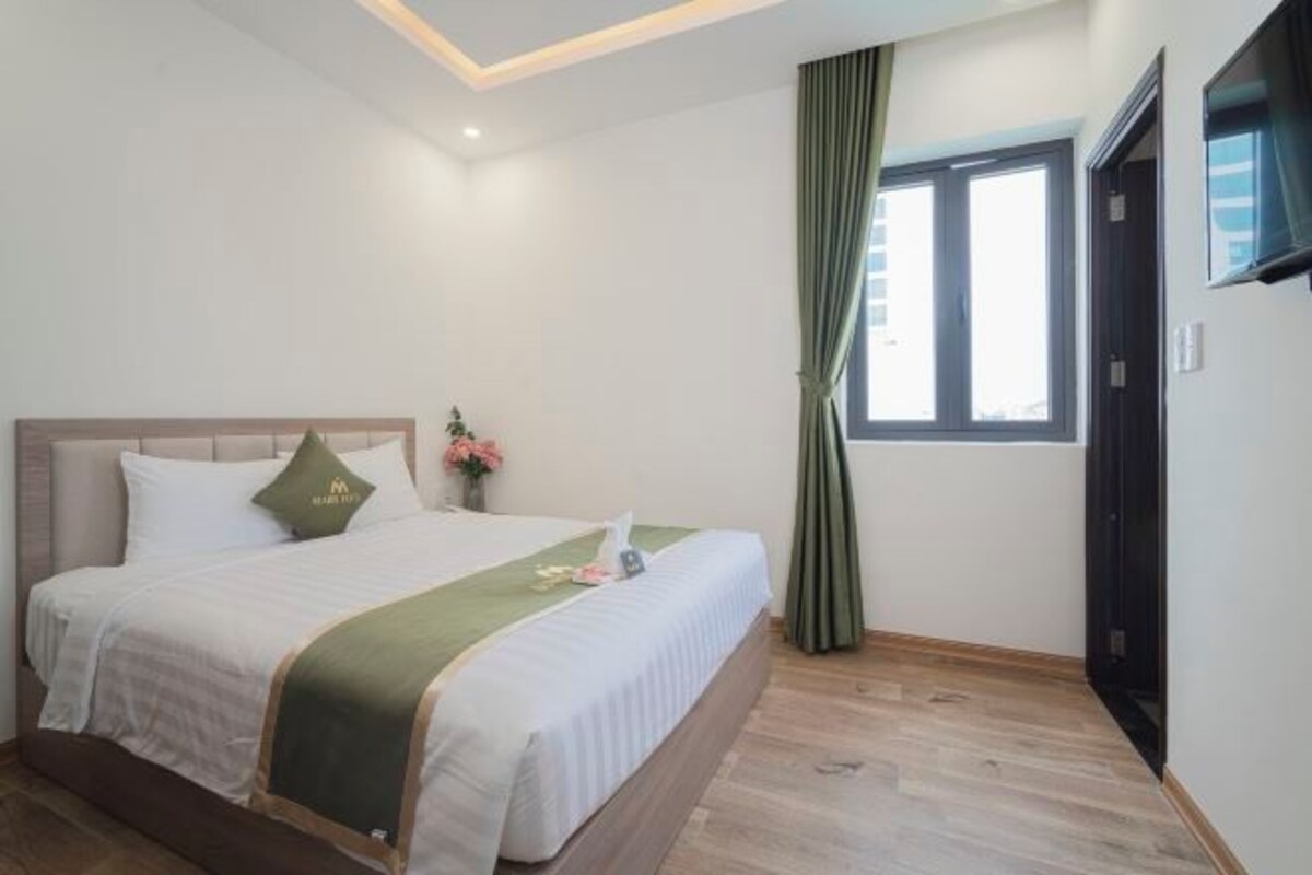 Habitación de hotel con una cama, una ventana con cortinas, una puerta y un televisor montado en la pared.