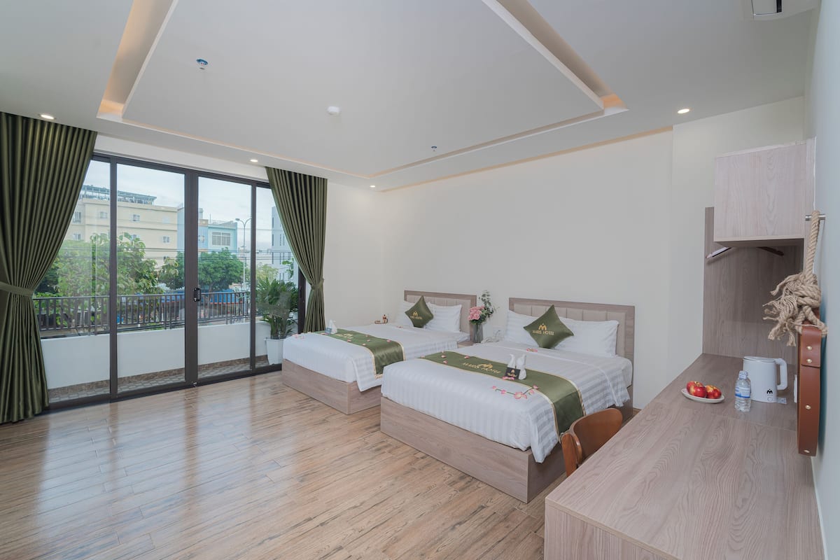 Ein Hotelzimmer mit zwei Betten, einem Balkon mit Vorhängen, Holzboden, einem Schreibtisch mit Kaffeemaschine und Blick auf Gebäude und Grünflächen.