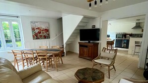 Un salon avec un canapé beige, une table à manger avec des chaises, un téléviseur et, au fond, un coin cuisine.