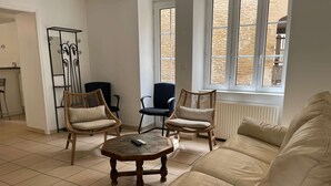 Un salon avec un canapé beige, deux fauteuils en osier, une petite table basse en bois et un radiateur près d’une fenêtre donnant sur un mur en pierre.