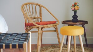 Une chaise en osier avec un coussin rouge, une petite table ronde en bois surmontée d’un vase de fleurs et un pouf jaune posé sur un tapis tissé.