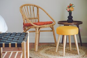 Une chaise en osier avec un coussin rouge, une petite table ronde en bois surmontée d’un vase de fleurs et un pouf jaune posé sur un tapis tissé.
