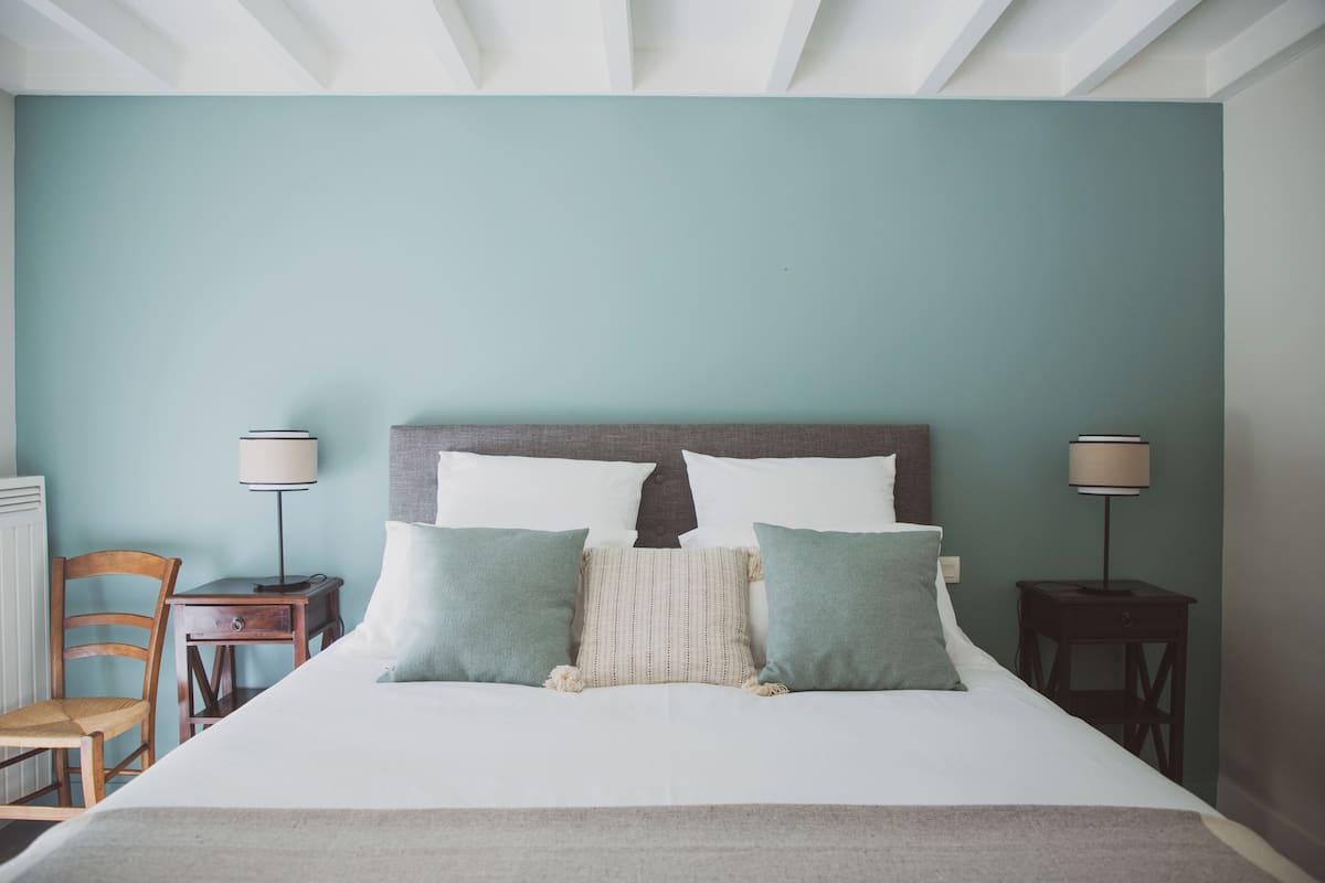 Een net opgemaakt bed met een houten hoofdbord, twee nachtkastjes met lampen, een stoel en een radiator.