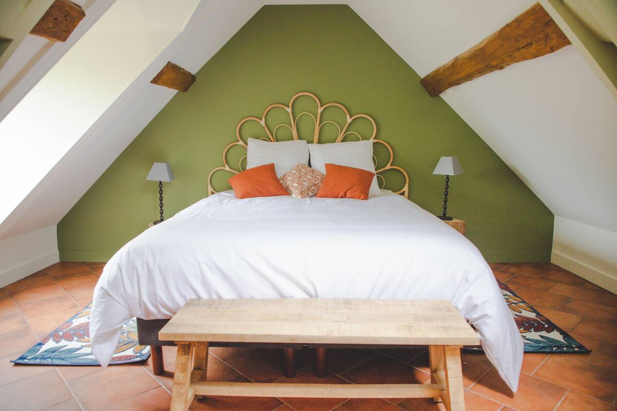 Een slaapkamer met een groene wand, een houten hoofdbord, een bed met wit en oranje beddengoed, twee nachtlampjes en een houten bankje.