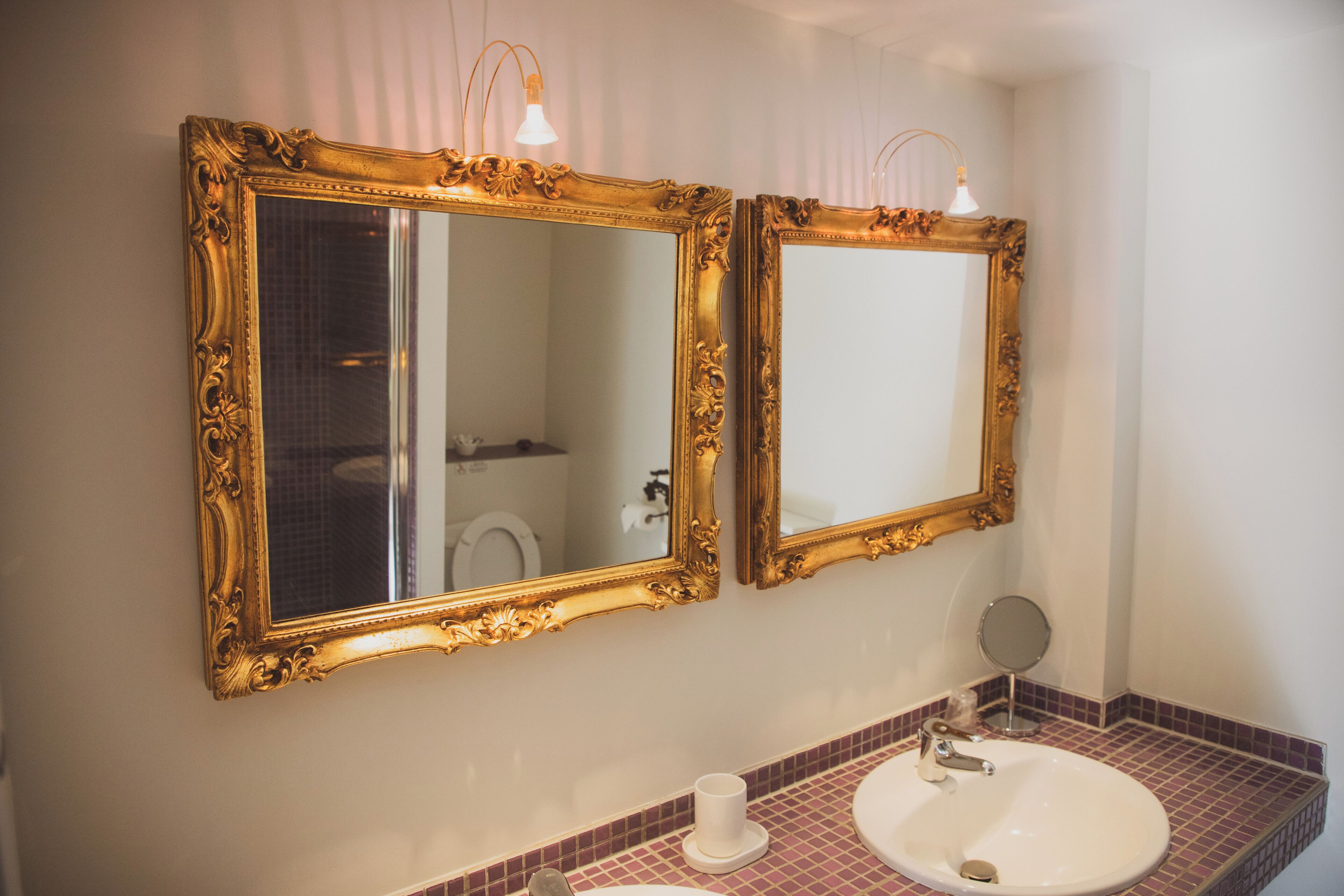 Een badkamer met twee sierlijke spiegel met gouden lijst, een witte wastafel en een kleine ronde spiegel.
