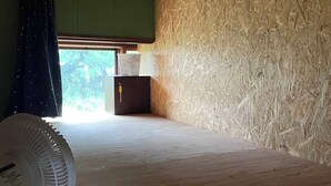 A room with a wooden ceiling, a fan on the floor, a window with a view of trees, and a door with a towel hanging on it.