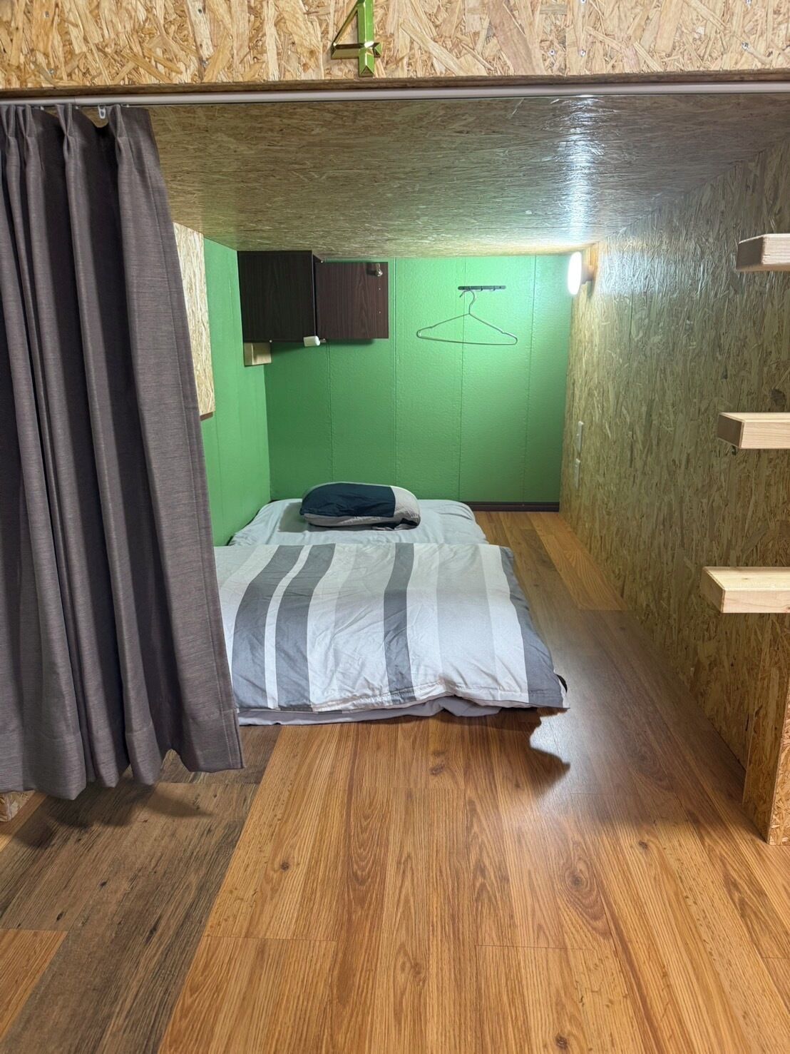 A small, compact room with a bunk bed, a green wall, a wooden floor, and a hanging clothes rack.