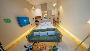Una habitación de hotel moderna con un sofá verde, una mesa de centro de madera con aperitivos y bebidas, una cama con almohadas blancas, un televisor y un espejo.