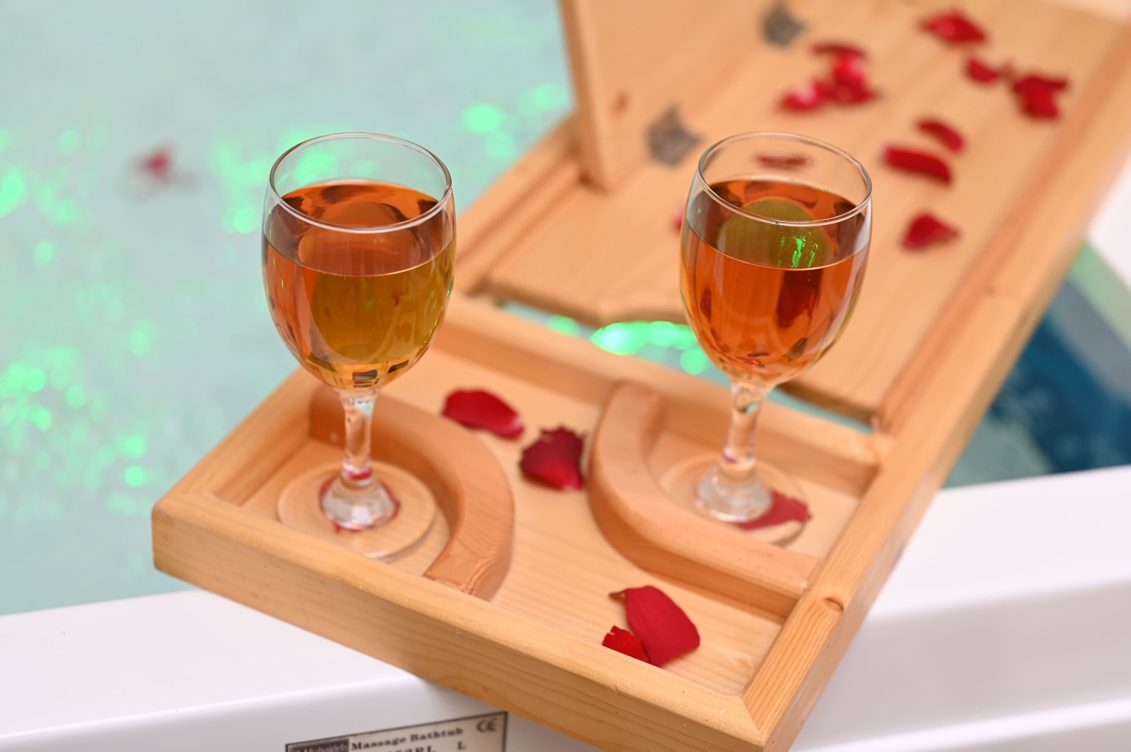 Two glasses of amber liquid on a wooden tray with rose petals, placed on a white surface.