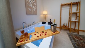 Un baño con una bañera, un estante de madera con una botella de champán, copas y pétalos de rosa.