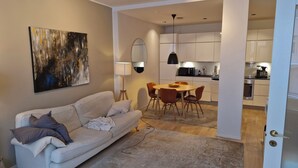 Een moderne woonkamer met een bank, een eettafel en stoelen. Een keuken met kasten en apparatuur is zichtbaar door een open doorgang.