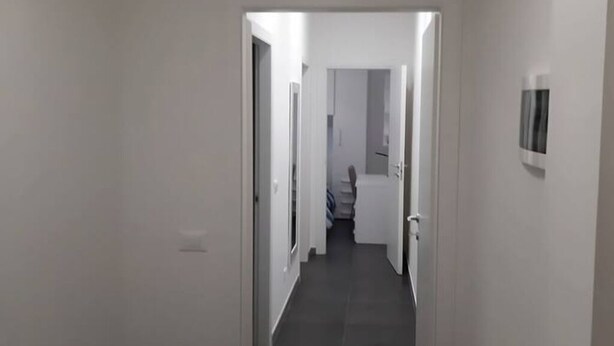 Kapea käytävä, jonka seinät ovat valkoiset ja lattia tumma, johtaa huoneeseen, jossa on sänky ja ovi.