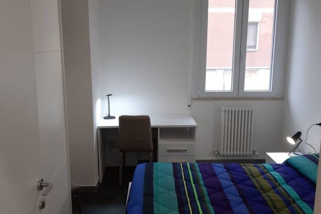 A bedroom with a bed, a desk, a chair, a radiator, and a window.
