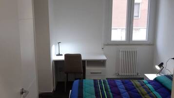 A bedroom with a bed, a desk, a chair, a radiator, and a window.