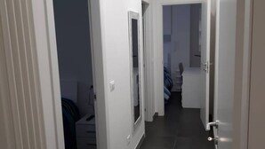 Kapea käytävä, jonka seinät ovat valkoiset ja jossa on laattalattia. Käytävä johtaa huoneeseen, jossa on sänky ja lipasto.
