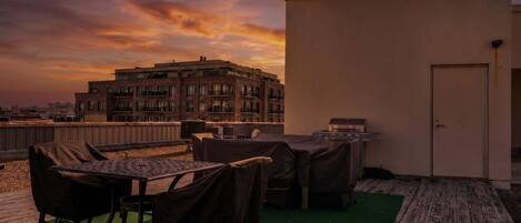 A rooftop terrace with a table and chairs, a barbecue grill, and a view of a building in the background.
