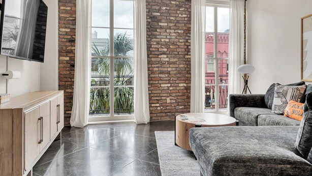 A modern living room with a brick wall, large windows with white curtains, a grey sofa, a wooden coffee table, and a TV mounted on the wall. - Chic 1BR in Warehouse District with. King bed, exposed brick, walk to FQ (New Orleans)