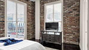 A bedroom with a brick wall, a large window, a bed, a TV on a wooden dresser, and a view of buildings outside. - Chic 1BR in Warehouse District with. King bed, exposed brick, walk to FQ (New Orleans)