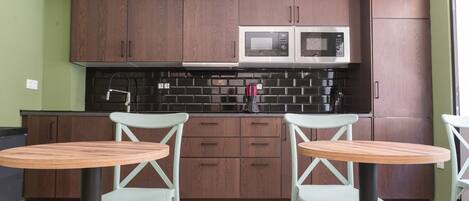 A compact kitchen with two built-in microwaves, a black backsplash, and wooden cabinets. There are two light green chairs around a small wooden table.
