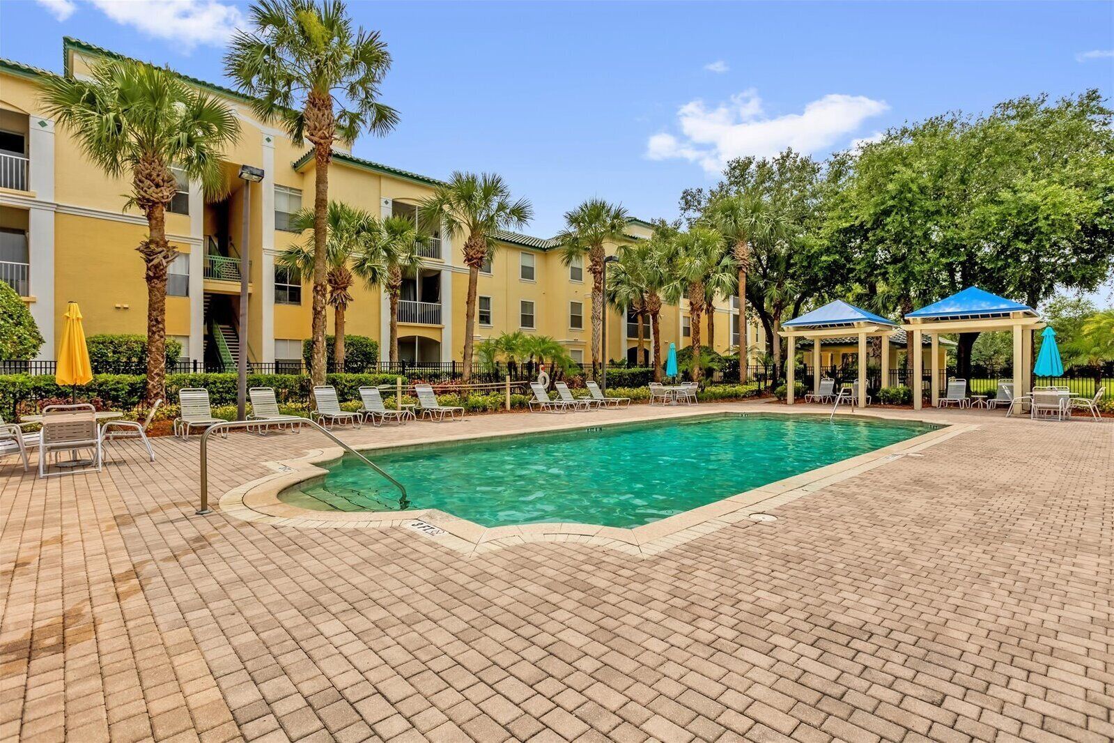 A pool area with lounge chairs and umbrellas, surrounded by palm trees and a building with balconies.