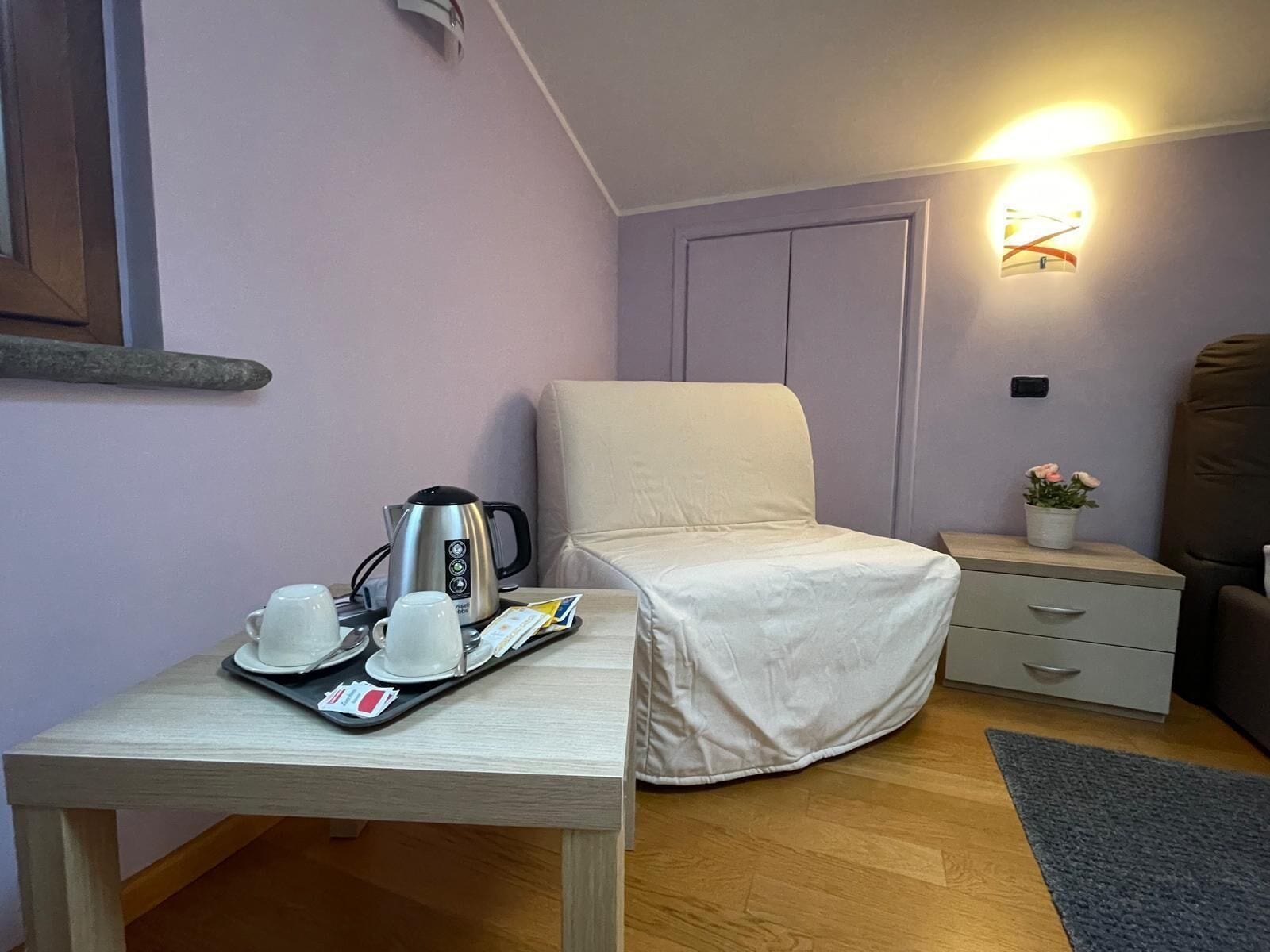 A small, cozy room with a single bed, a bedside table with a tray of breakfast items, a wall-mounted light, and a small potted plant.