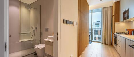 A modern hotel room with a glass shower, a toilet, a sink, a wooden floor, a kitchen area with a stove, and a view of the city through large windows.