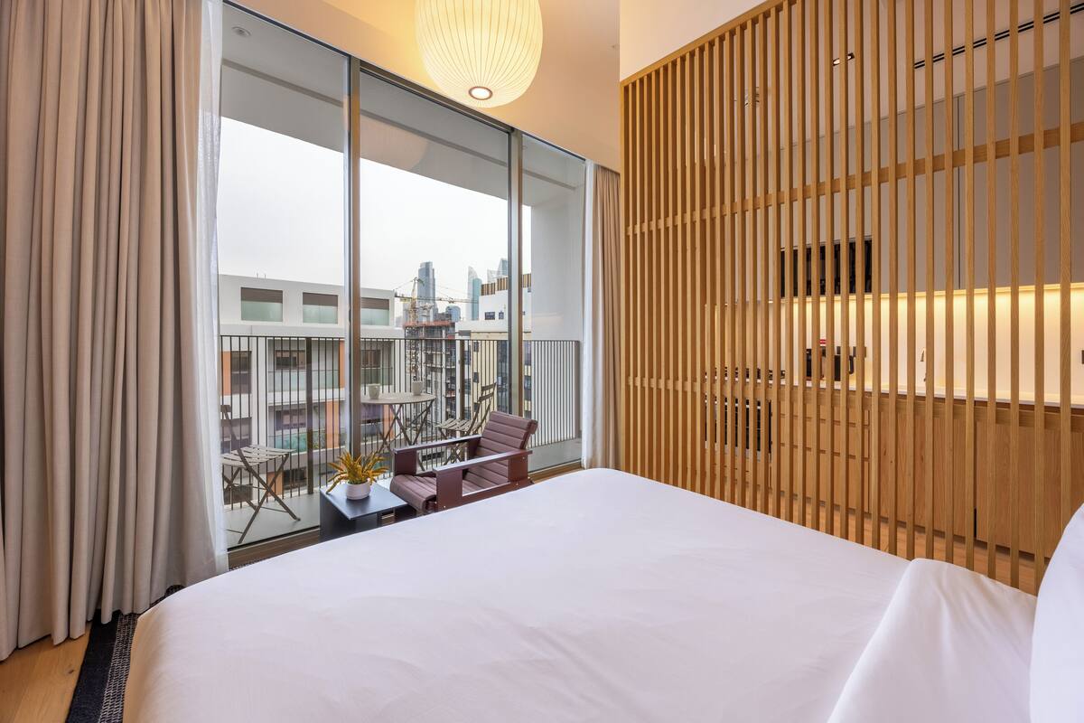 A modern hotel room with a large bed, a balcony with a table and chairs, and a view of the cityscape.