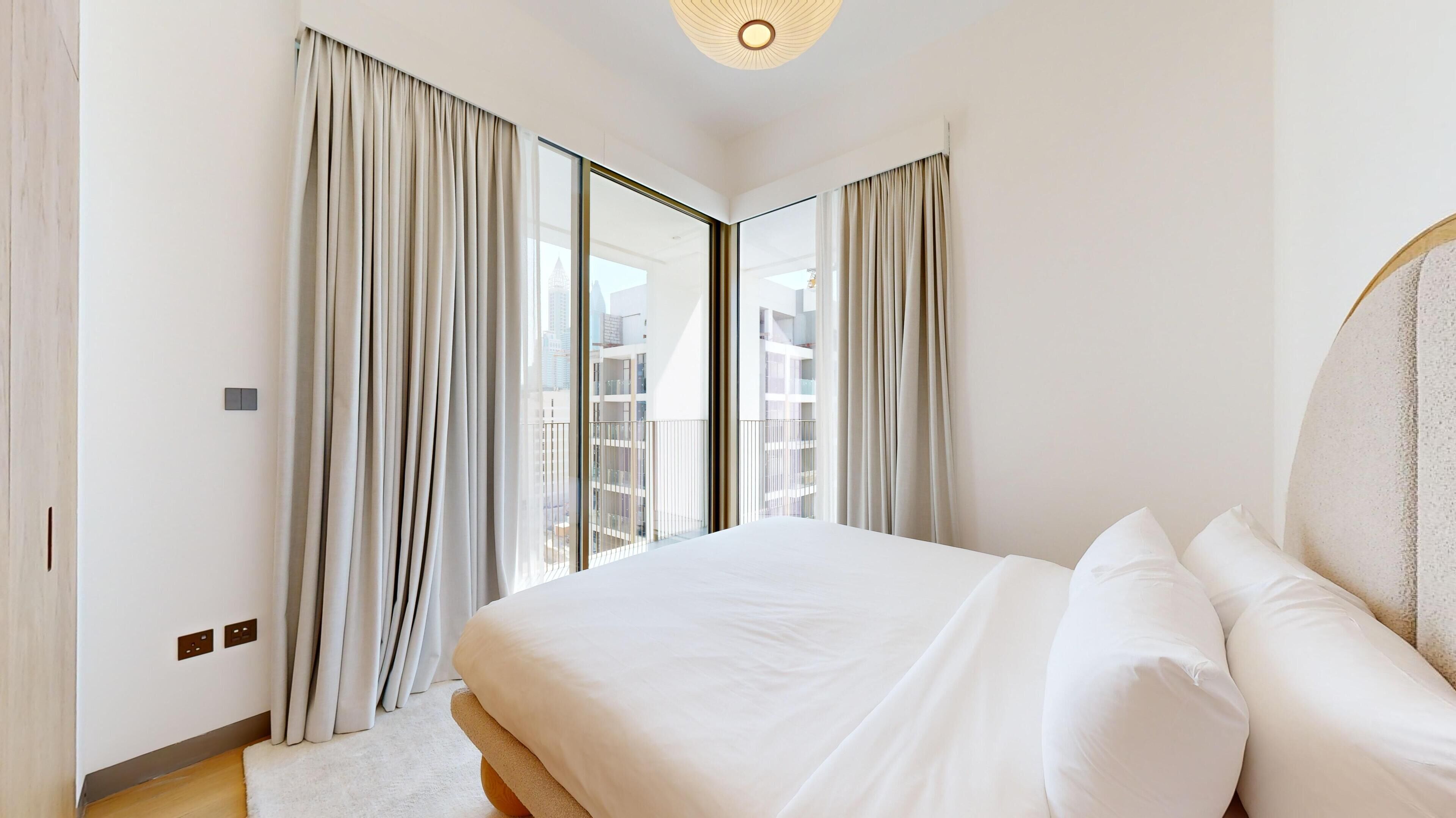 Une chambre d’hôtel moderne avec un grand lit, deux fenêtres avec rideaux, une vue sur le paysage urbain et un luminaire au plafond.
