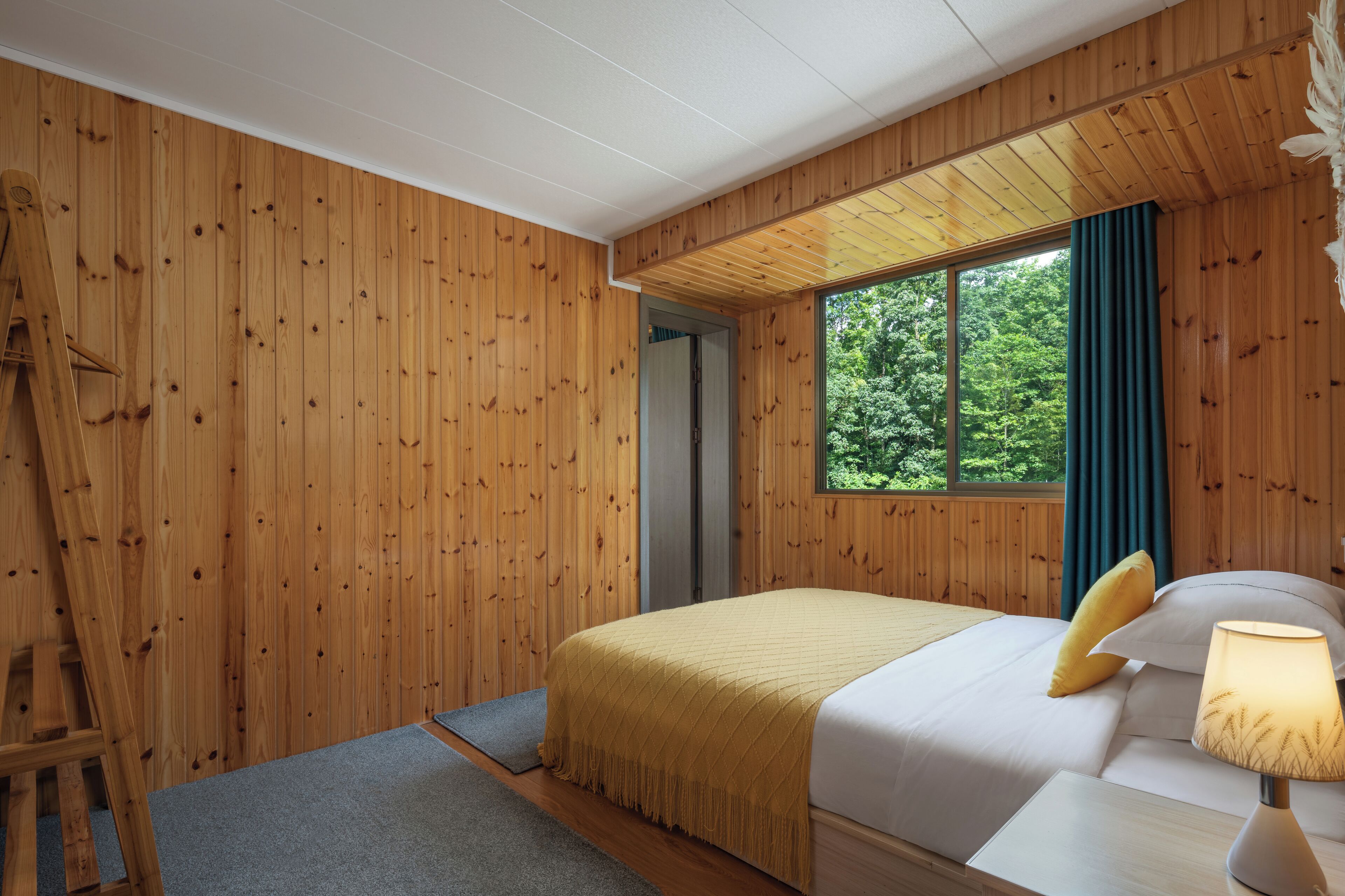 A wooden-paneled room with a bed, a nightstand, a lamp, and a window with a view of greenery.