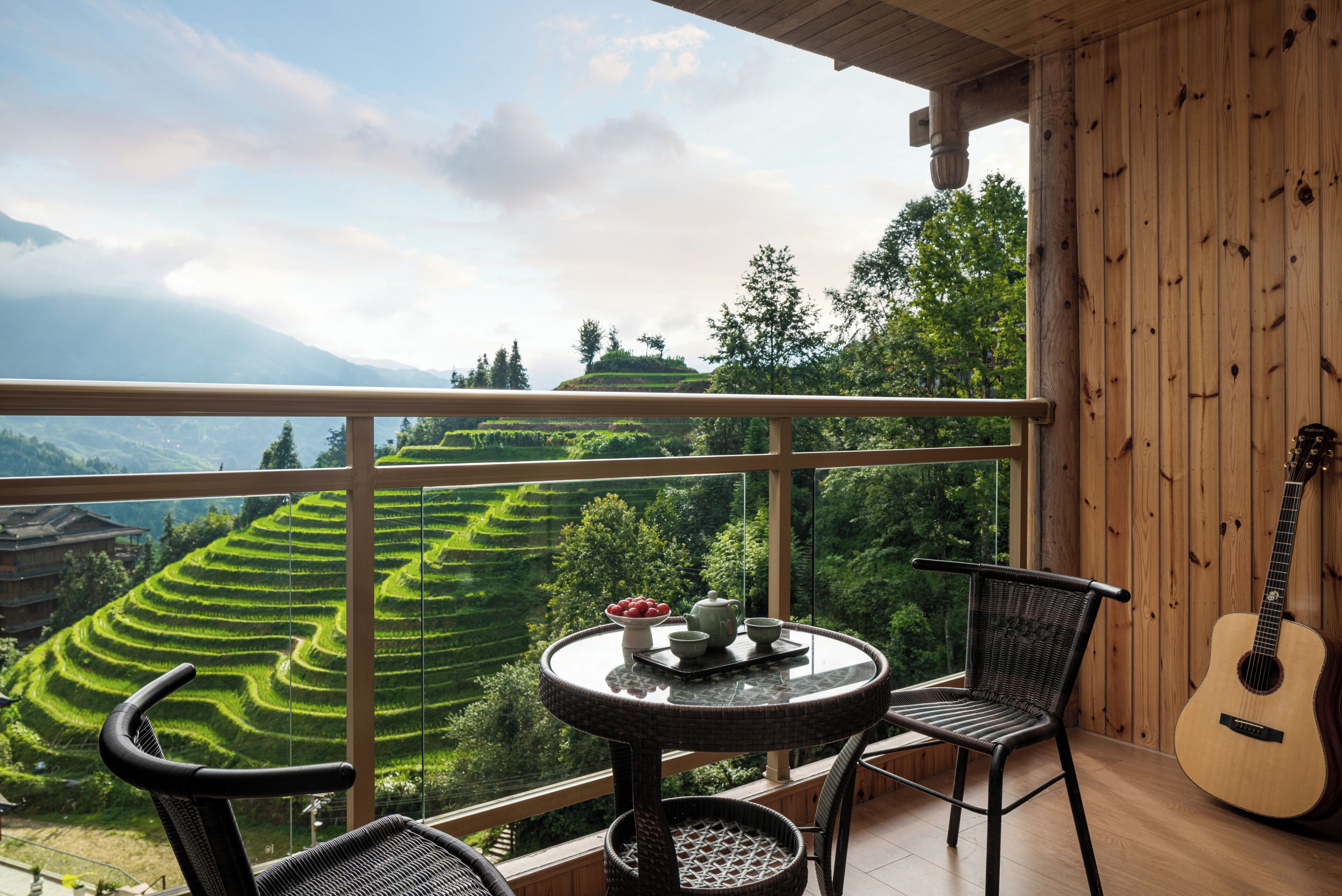 A balcony with a table, chairs, and a guitar, overlooking terraced fields and a mountainous landscape.