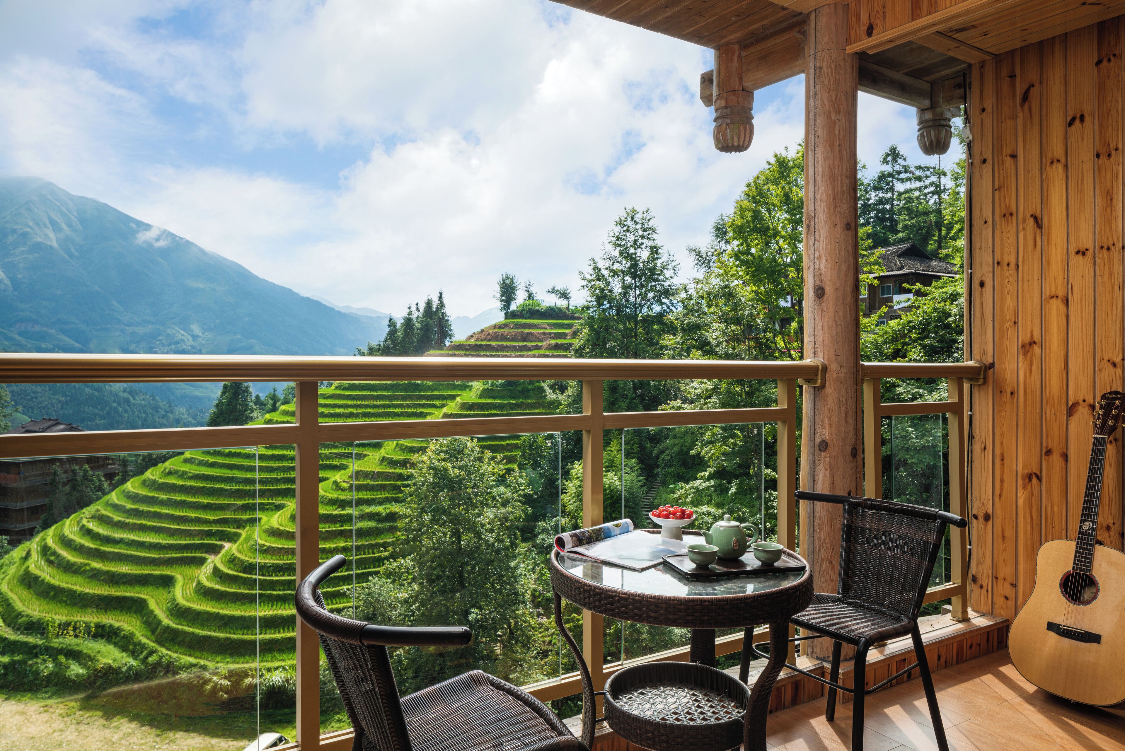 A wooden balcony with a table, chairs, and a guitar, overlooking terraced fields and mountains.