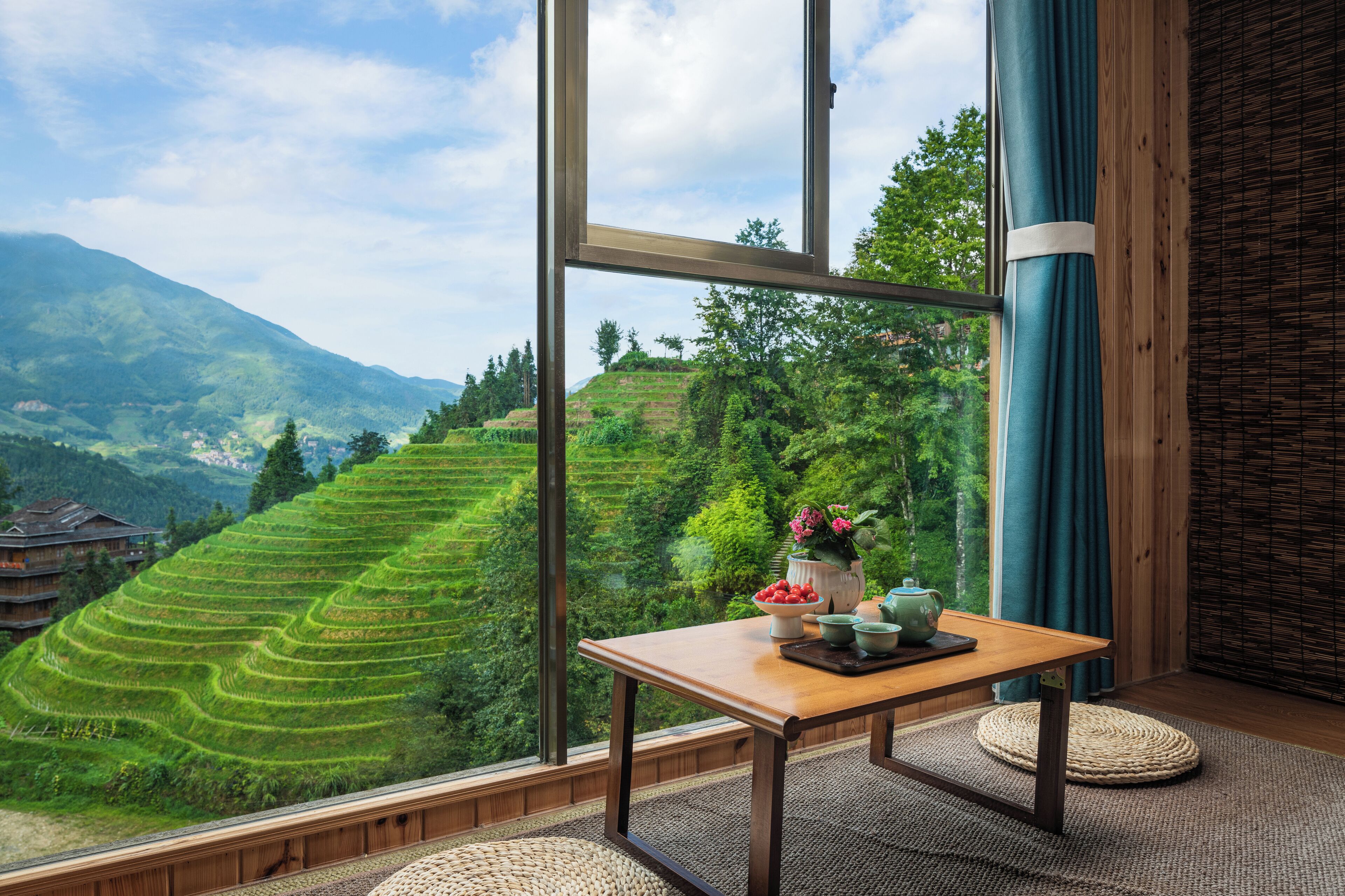 A room with a large window offering a view of terraced fields and mountains.