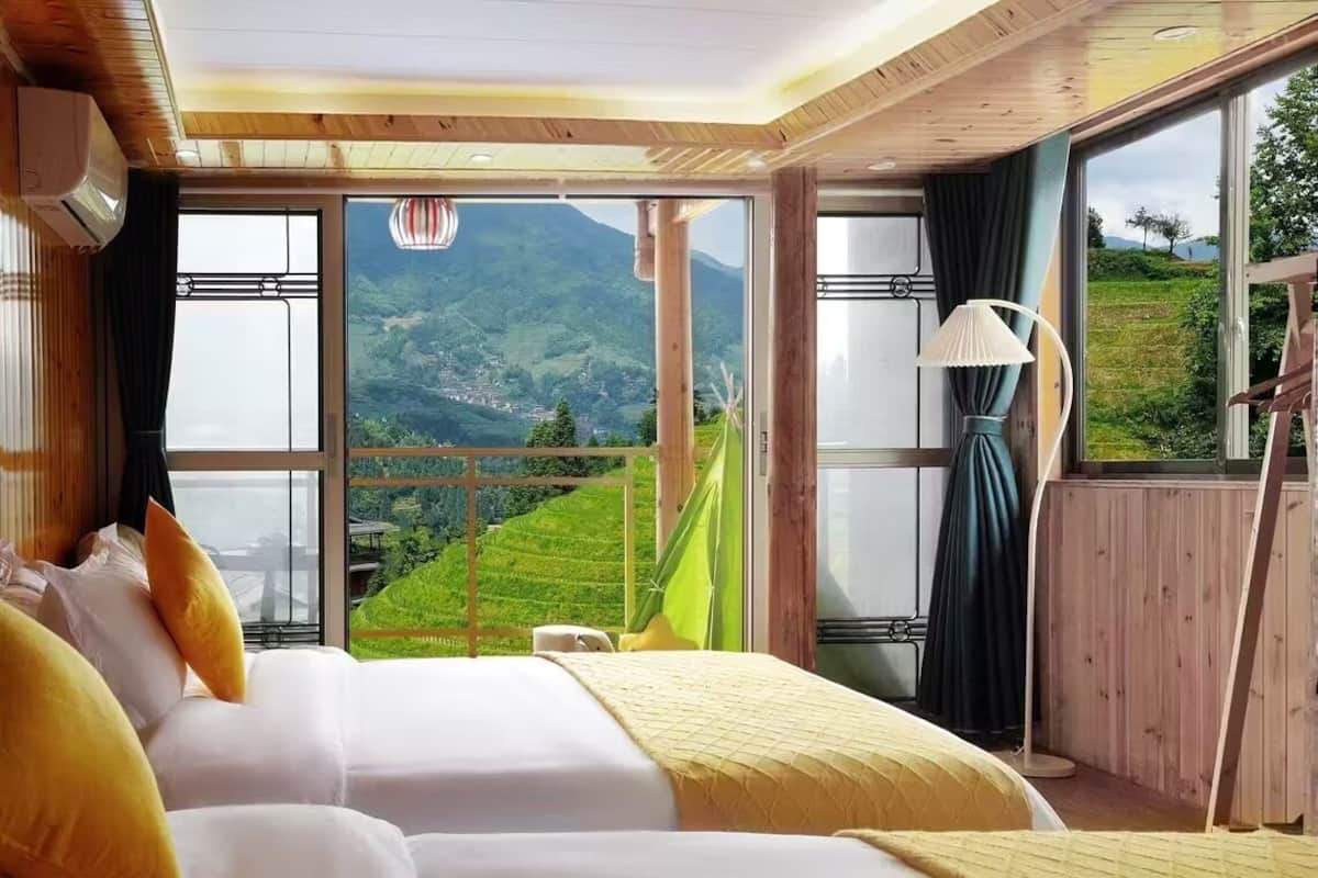 A hotel room with two beds, large windows offering a view of a mountainous landscape, a hanging chair, and a lamp.