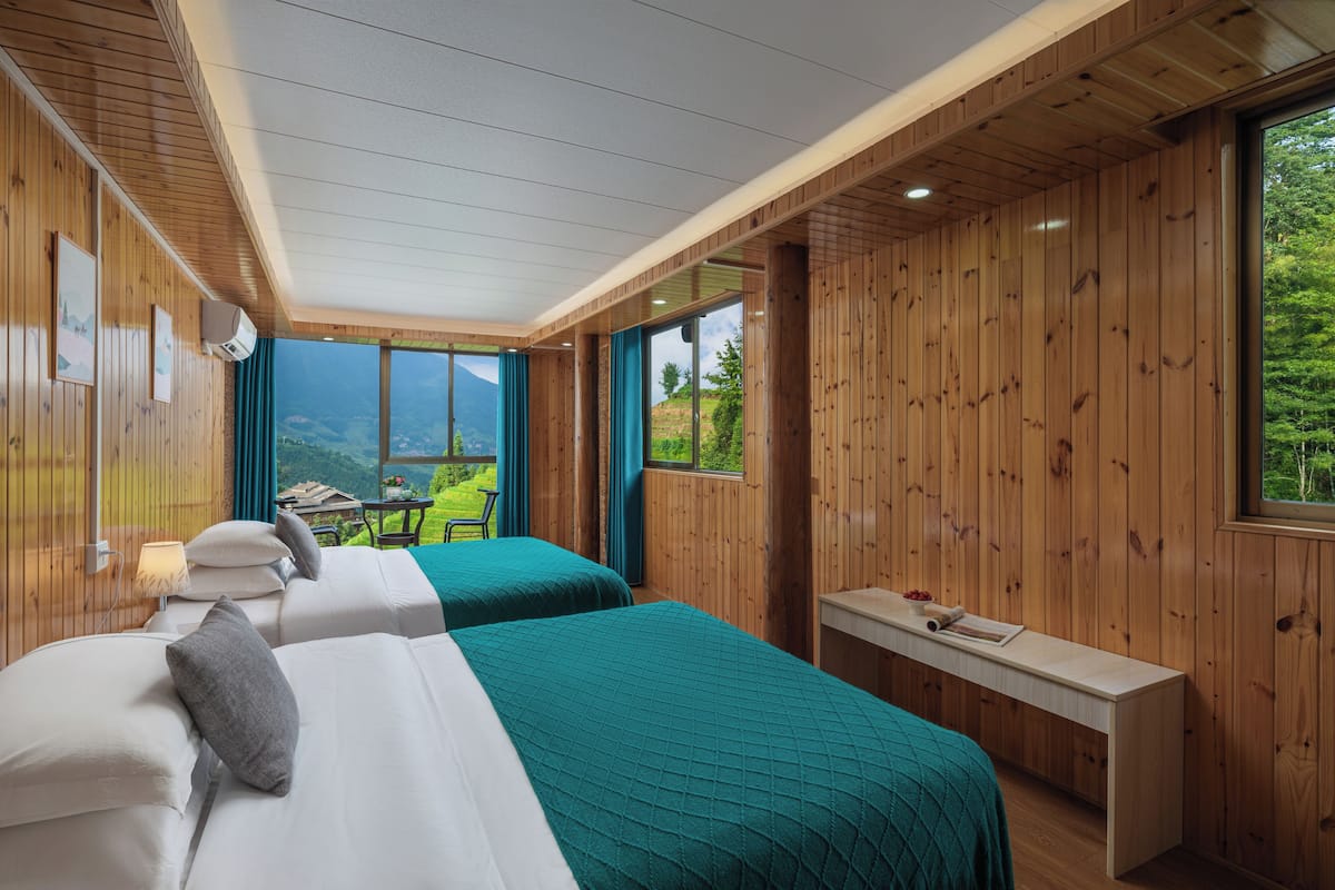 A bedroom with two beds, a wooden ceiling, a window with a view of greenery, and a small table with a lamp.