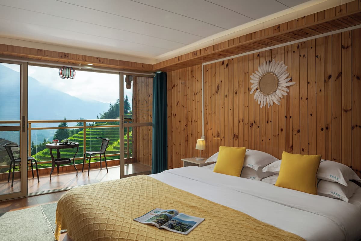 A bedroom with a large bed, wooden walls, a view of a mountainous landscape, and a dining area with a table and chairs.
