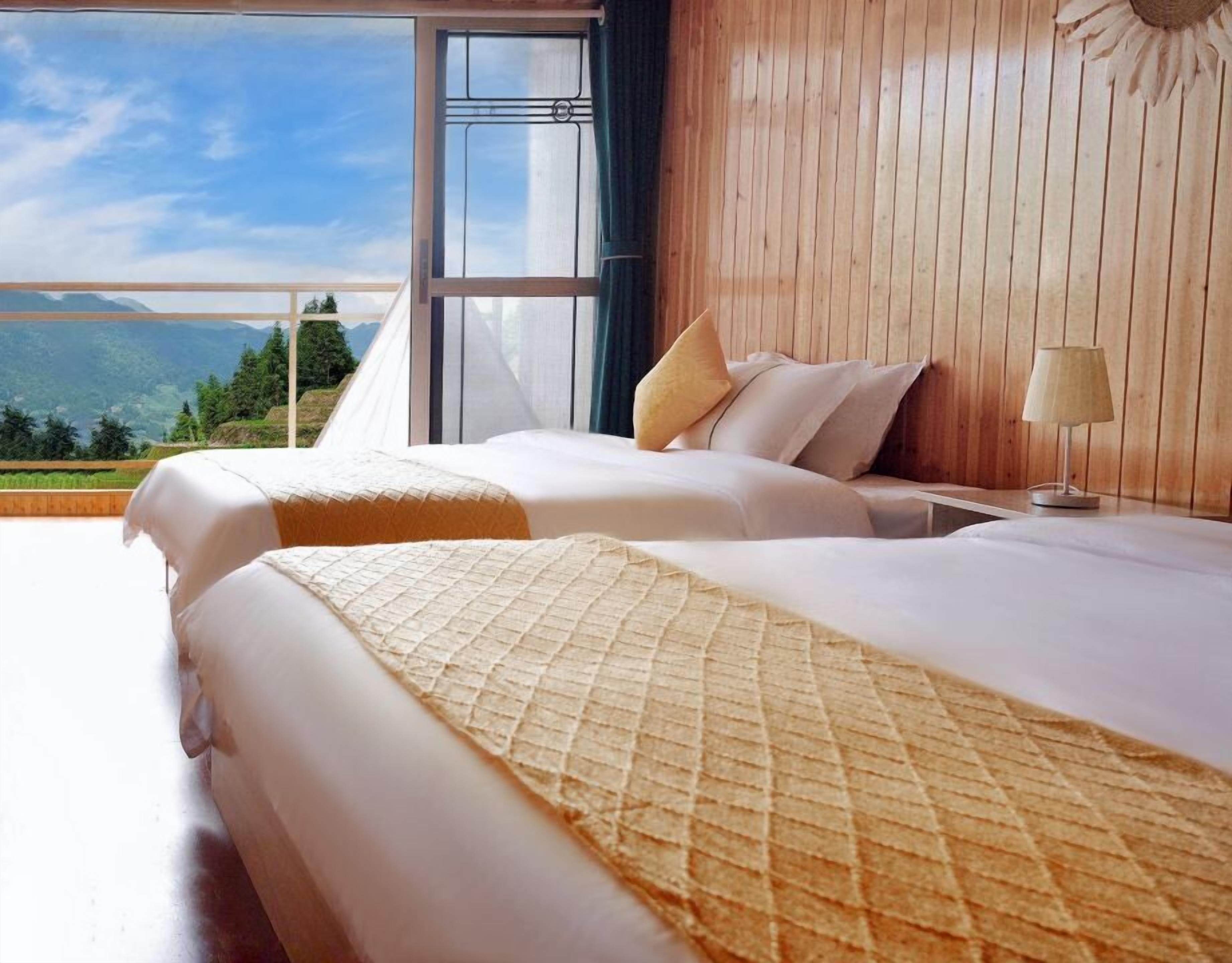 A hotel room with two beds, a large window with a view of mountains, and wooden paneling on the walls.