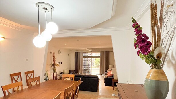 A spacious dining area with a long wooden table and chairs, a modern ceiling light fixture, and a vase with flowers on a wooden cabinet. - Parry Suite (Weija)