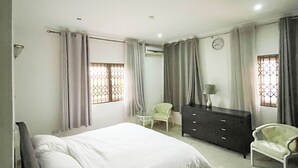 A bedroom with a bed, a dresser, two chairs, and a window with curtains. - Parry Suite (Weija)