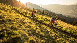 Two cyclists on a grassy mountain trail with a scenic view of distant hills and a small building in the background. - Mountain & Golf Resort Westendorf (Westendorf)