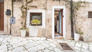 A stone building with a white door, a window with a metal frame, and a potted plant in front. - Vico di Ostuni Apartments by Rentbeat (Ostuni)