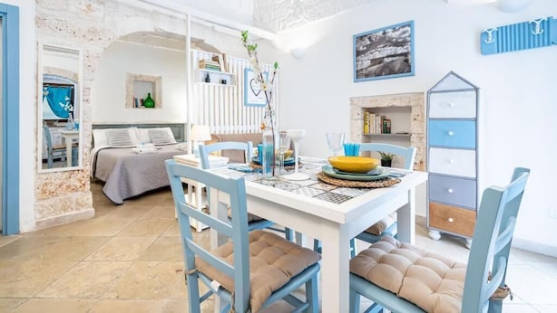 A bright, open-plan living area with a dining table set for two, a cozy bed, and a small kitchen area. - Vico di Ostuni Apartments by Rentbeat (Ostuni)
