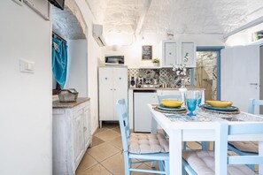 A compact kitchen with white cabinets, a microwave, and a small refrigerator. A dining table with blue chairs and yellow bowls is set for two. - Vico di Ostuni Apartments by Rentbeat (Ostuni)
