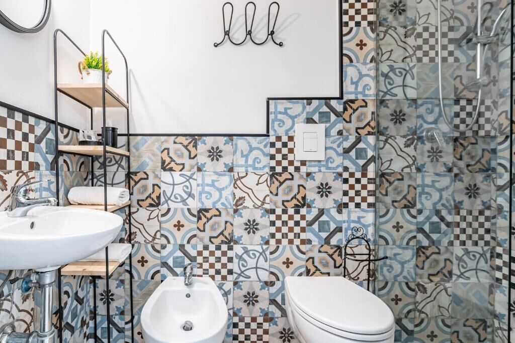 A bathroom with a white sink, a bidet, and a toilet. There is a shelf with a plant and a towel. The walls are decorated with patterned tiles.