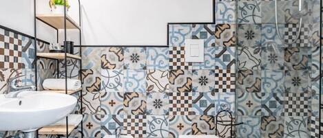 A bathroom with a white sink, a bidet, and a toilet. There is a shelf with a plant and a towel. The walls are decorated with patterned tiles.